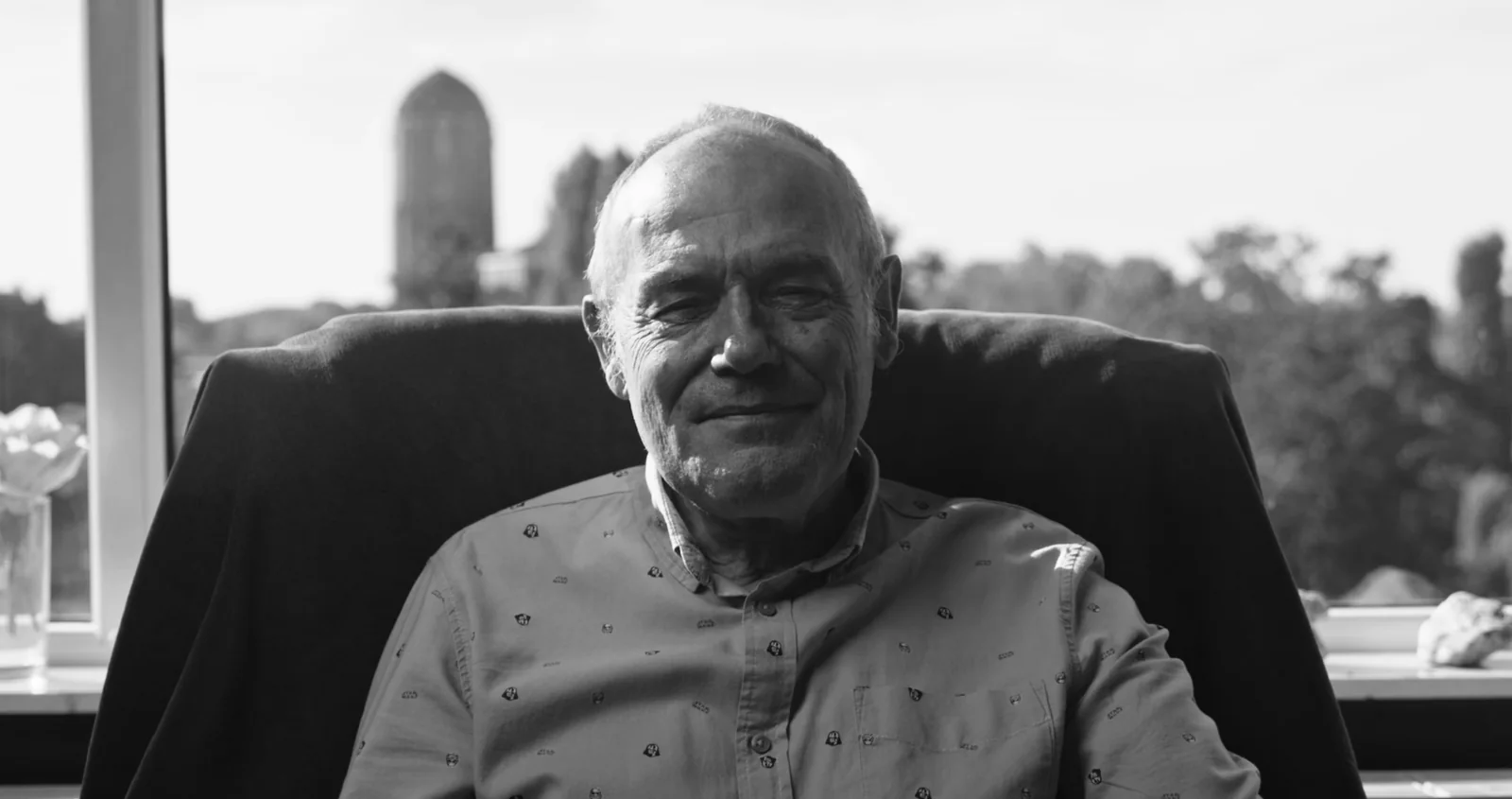 Wilbert, an older man smiling with his eyes closed, seated by a window.