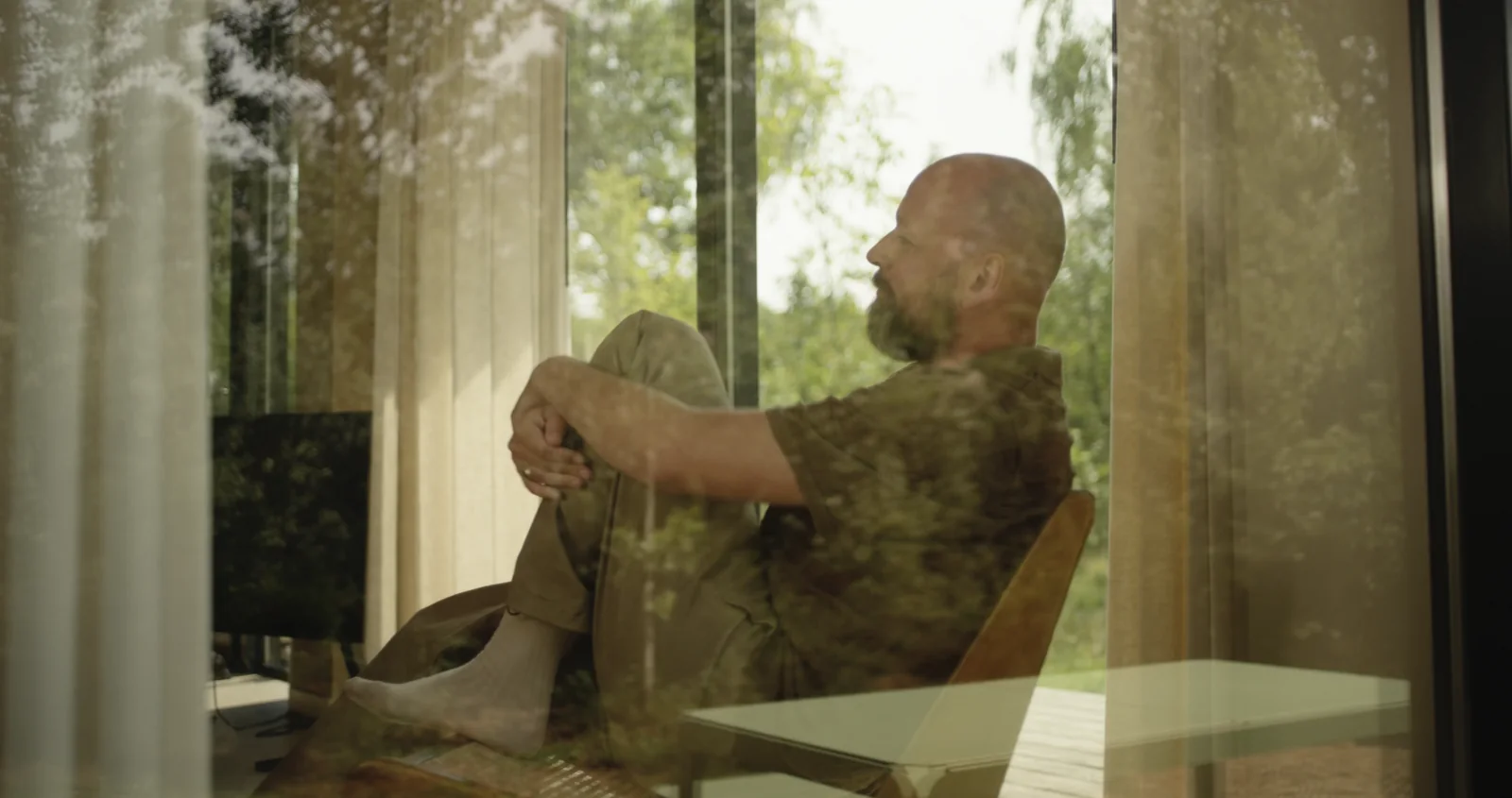 A man sitting by a tall window, looking out at the trees.